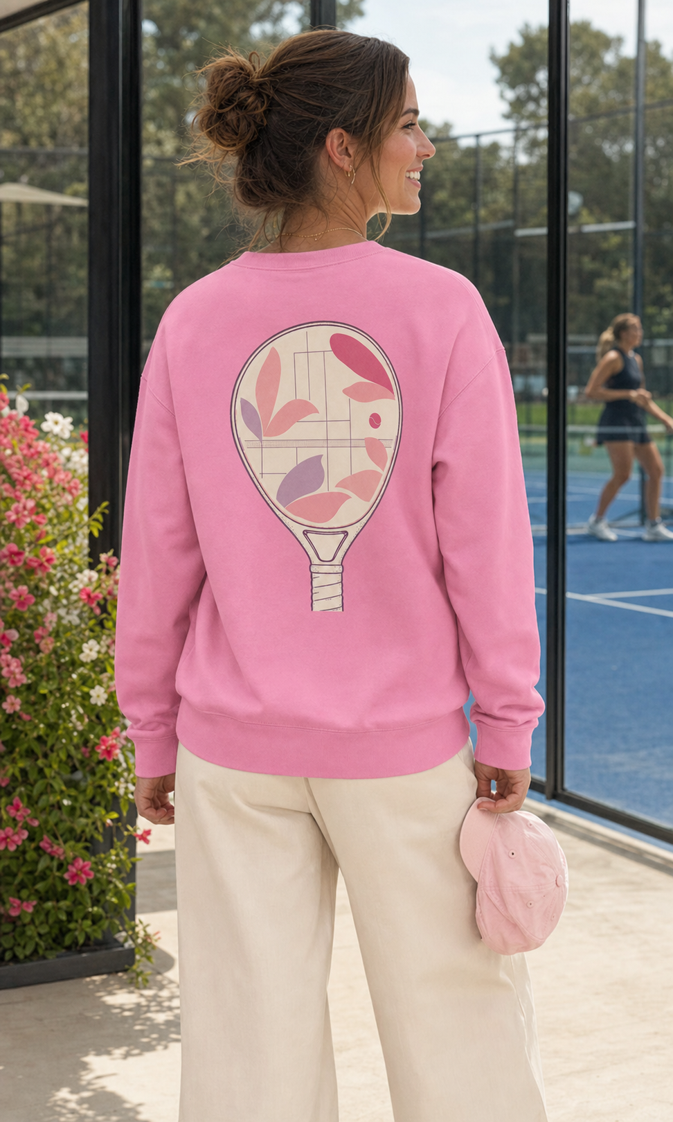 A woman on an outdoor padel court wearing a pink crewneck sweatshirt with large floral padel racket-window artwork on the back, paired with cream trousers.
