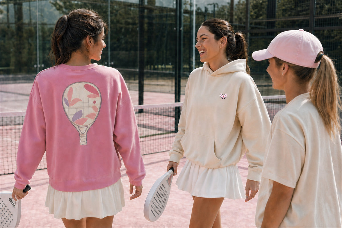 Three women on a padel court seen from behind, wearing After Padel Club apparel: a pink racket-print crewneck sweatshirt, a cream oversized hoodie with an embroidered racket logo, and a pink baseball cap with a cream top.